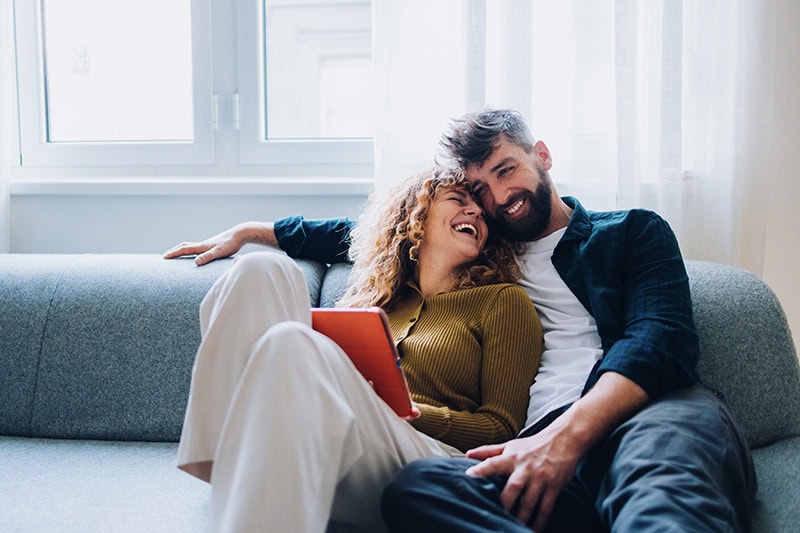 Smiling couple enjoying leisure time together at home, sitting on a cozy sofa by a window. Their joyful expressions highlight their close relationship and happiness while sharing a moment of connection.